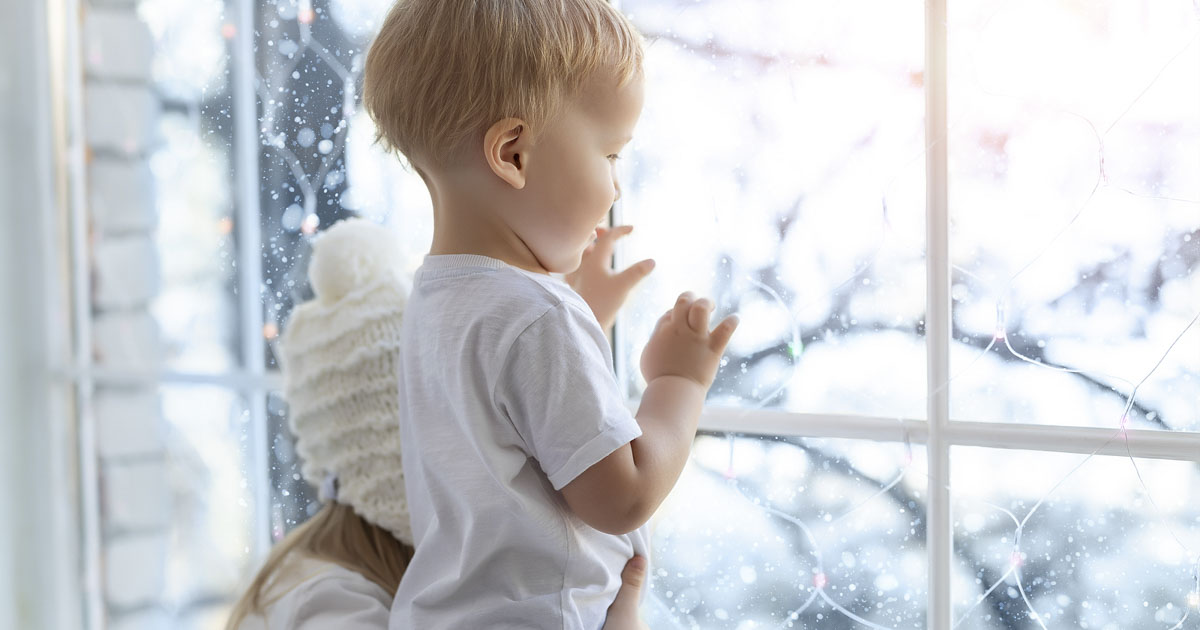 Two siblings stand by a window looking out at snow falling, with a cozy indoor setting.